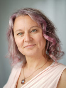 A woman with pink hair, a pink shirt and a mischievous smile